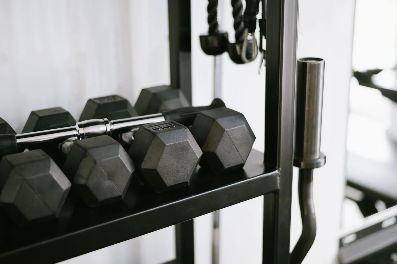 Haltères hexagonaux noirs rangés sur un rack, avec des poignées de poulie suspendues au-dessus