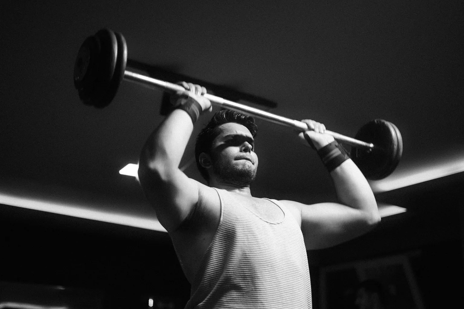 Photo en noir et blanc d'un homme poussant une barre chargée au-dessus de sa tête dans une salle de sport